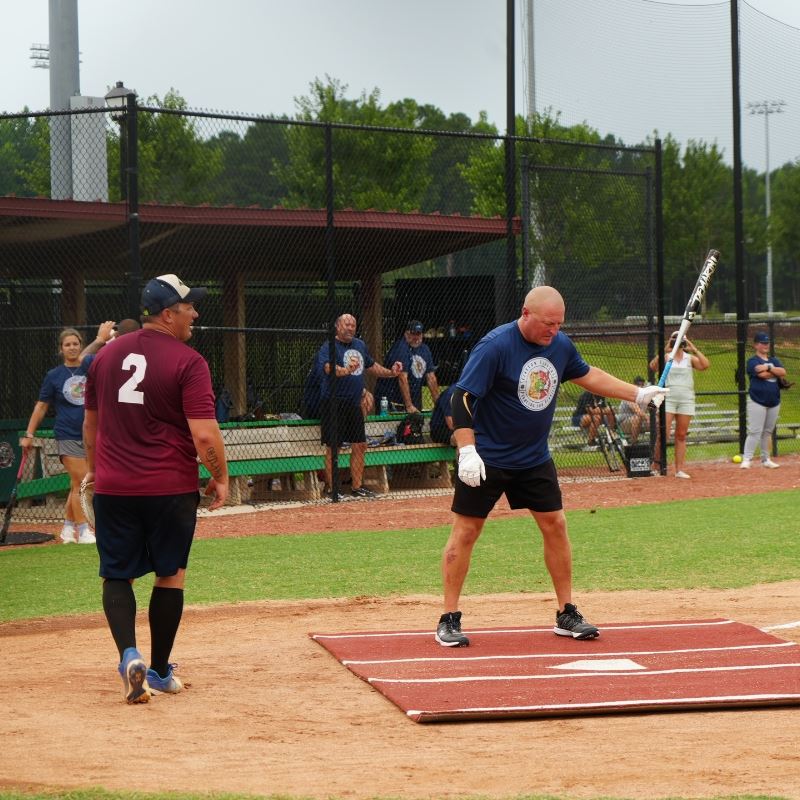 Officer in blue tshirt gets ready to bat at home plate with fire fighter in red t-shirt is behind